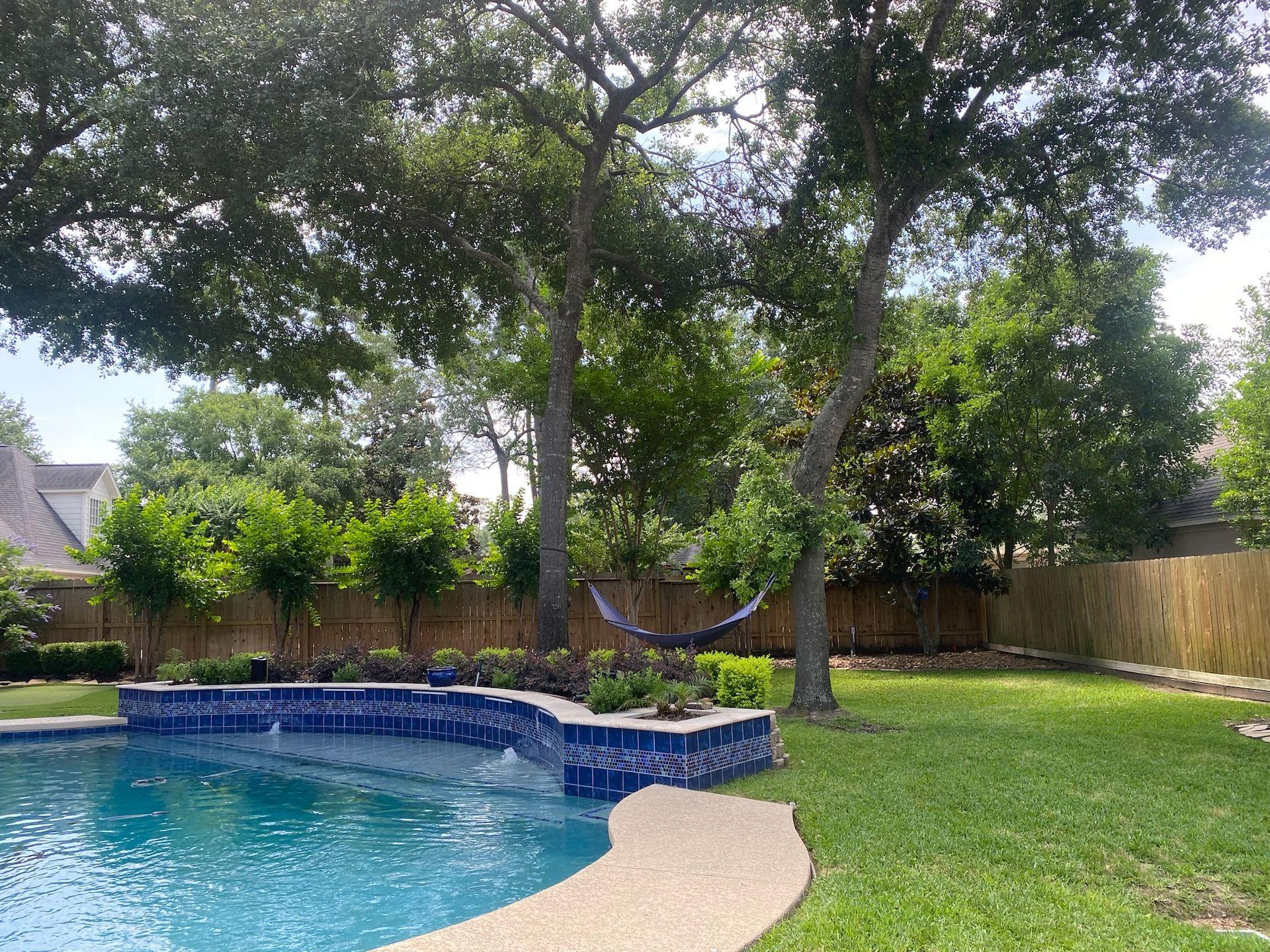 Stunning Houston backyard oasis with professional landscaping, including a crystal-clear swimming pool, lush green lawn, and mature trees for natural shade. This beautifully designed outdoor space features an in-ground trampoline, making it perfect for families seeking safe and fun backyard activities. The landscape includes meticulously maintained flower beds, a cozy hammock for relaxation, and a wooden privacy fence. Ideal for those searching for top Houston landscaping services, backyard pool designs, and in-ground trampoline installations. Transform your outdoor space with expert Houston landscapers who specialize in creating luxurious, family-friendly environments. Explore the best landscaping ideas for Houston, TX, and discover how to integrate a pool, lawn, and in-ground trampoline seamlessly into your backyard design.