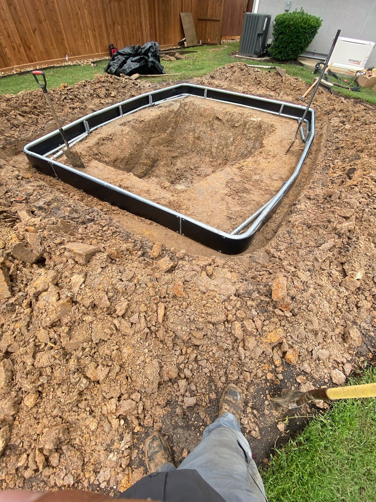 Excavation site for backyard landscaping project with dug-out area, soil, and nearby wheelbarrow and shovel, surrounded by grassy lawn and wooden fence