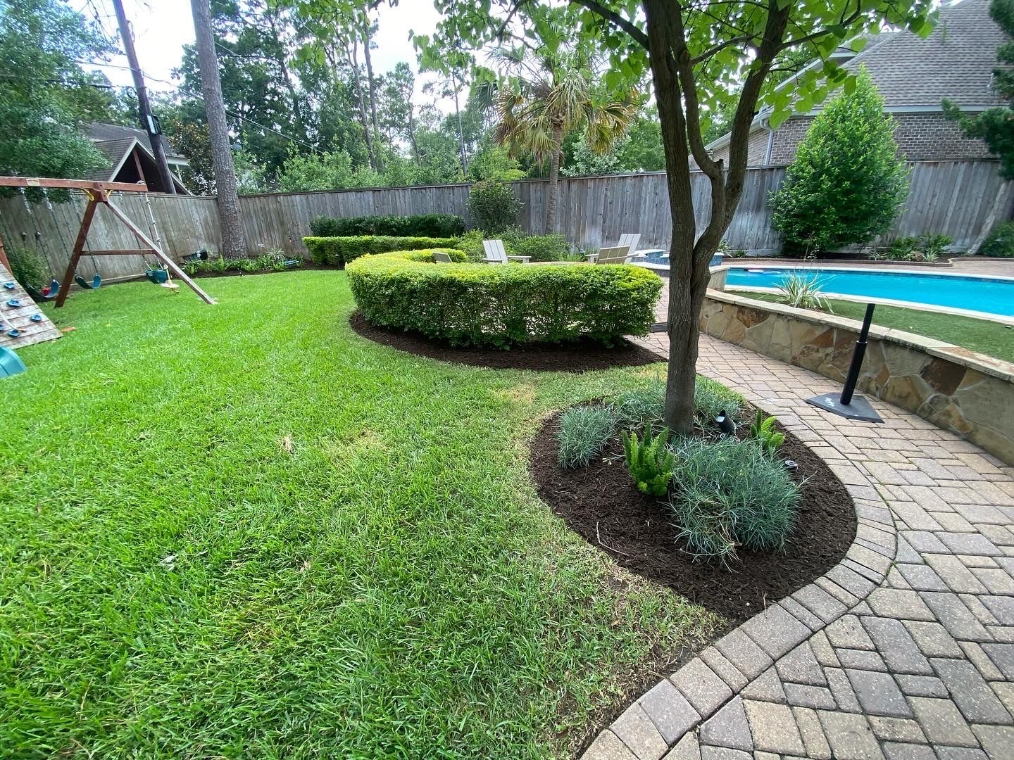 Beautifully landscaped backyard with lush green grass, neatly trimmed hedges, and a tree with mulch bed. Features include a swing set, a paved walkway, and a swimming pool surrounded by a wooden fence.