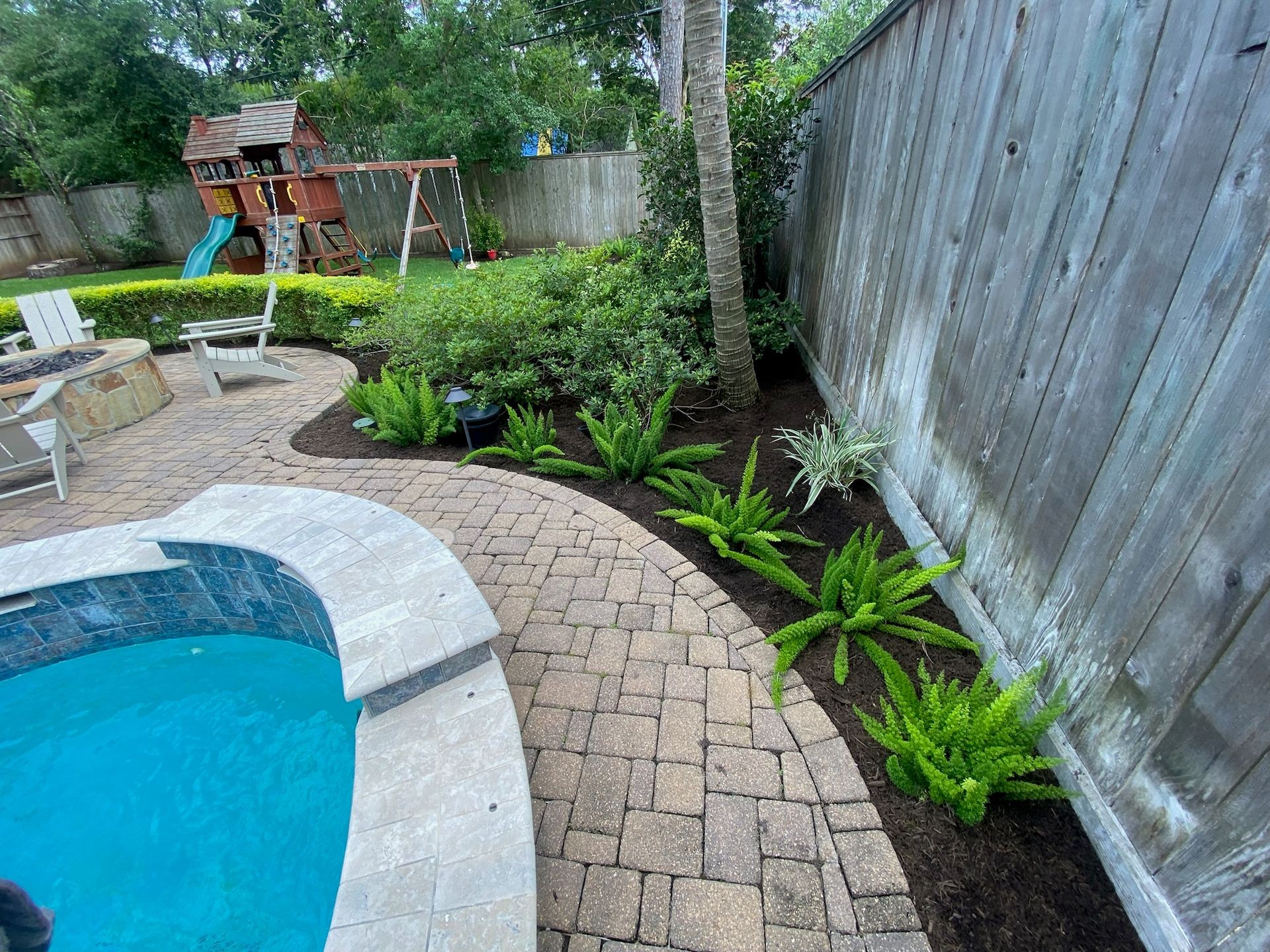 Backyard with stone pool, paved patio, fire pit, landscaped garden with ferns and other plants, children's playset, wooden fence